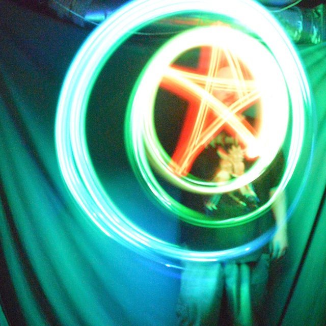 A long-exposure photograph: rings of teal light around an orange five-pointed star drawn in the air, with a blurred figure standing under it.